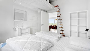 Quarto moderno com cama grande, roupeiro embutido, banheiro com espelho e pia, e vista para o exterior.