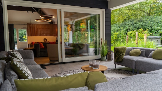 A modern living room with grey sofas, a coffee table, and a sliding glass door leading to a kitchen and garden. - Villa in Brabant With Spa and Sauna (Oirschot)
