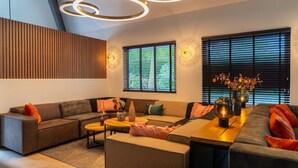 A modern living room with a sectional sofa, a wooden accent wall, and a large window with blinds. - Villa in Brabant With Spa and Sauna (Oirschot)