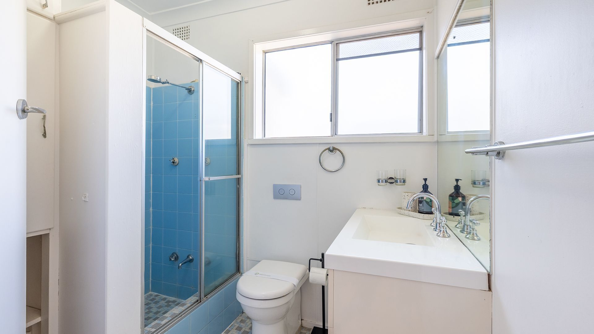 A bathroom with a glass shower enclosure, a toilet, a sink with a mirror, and a window above the sink.