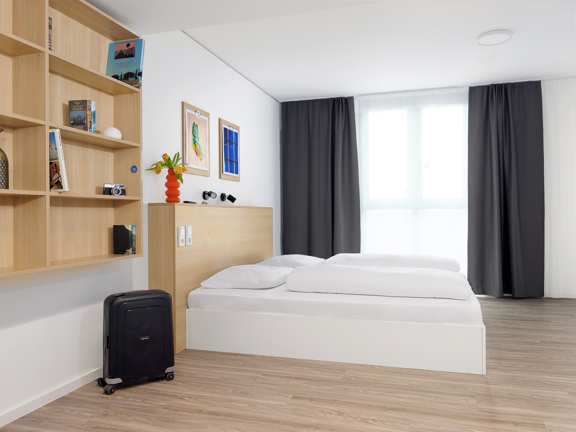 A hotel room with a bed, a wooden shelf with books and a camera, a suitcase, and a window with curtains.