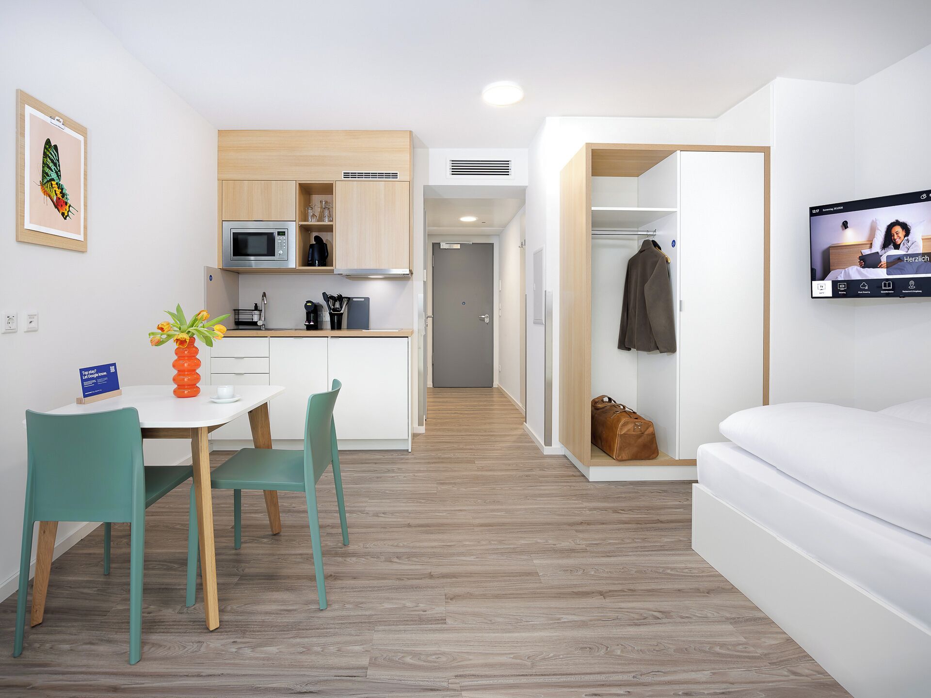 A modern hotel room with a kitchenette, a dining area with a table and chairs, a TV mounted on the wall, and a walk-in closet.
