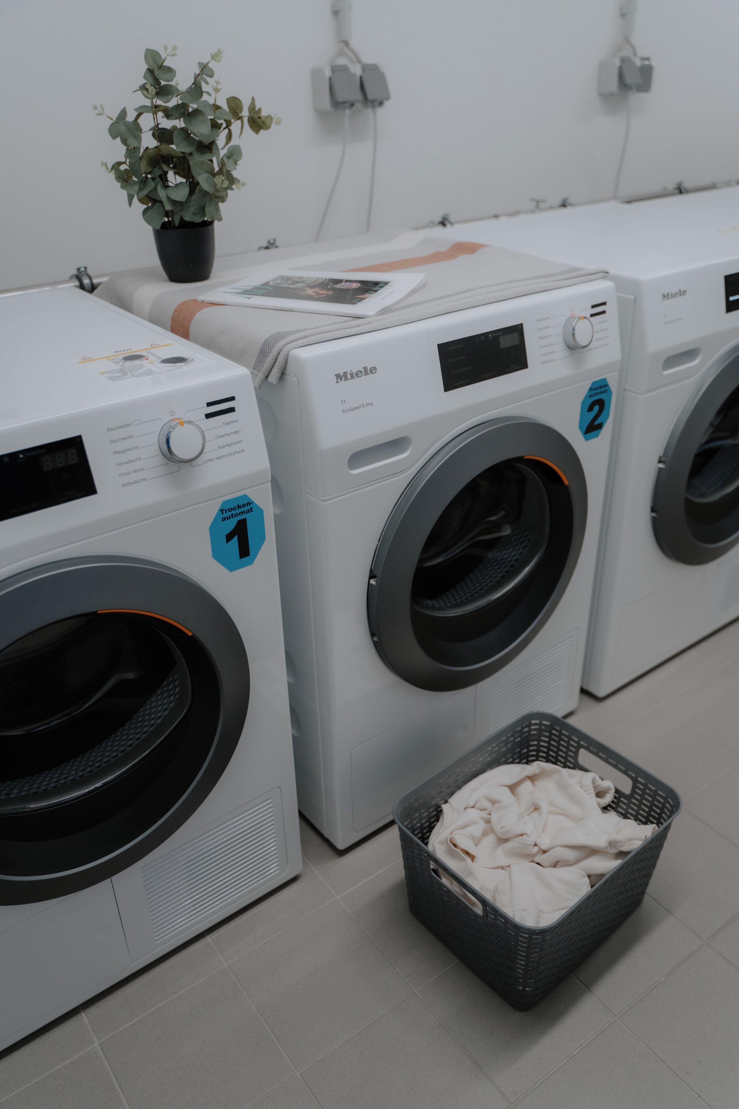 A row of white front-loading washing machines with a basket of clothes in front.