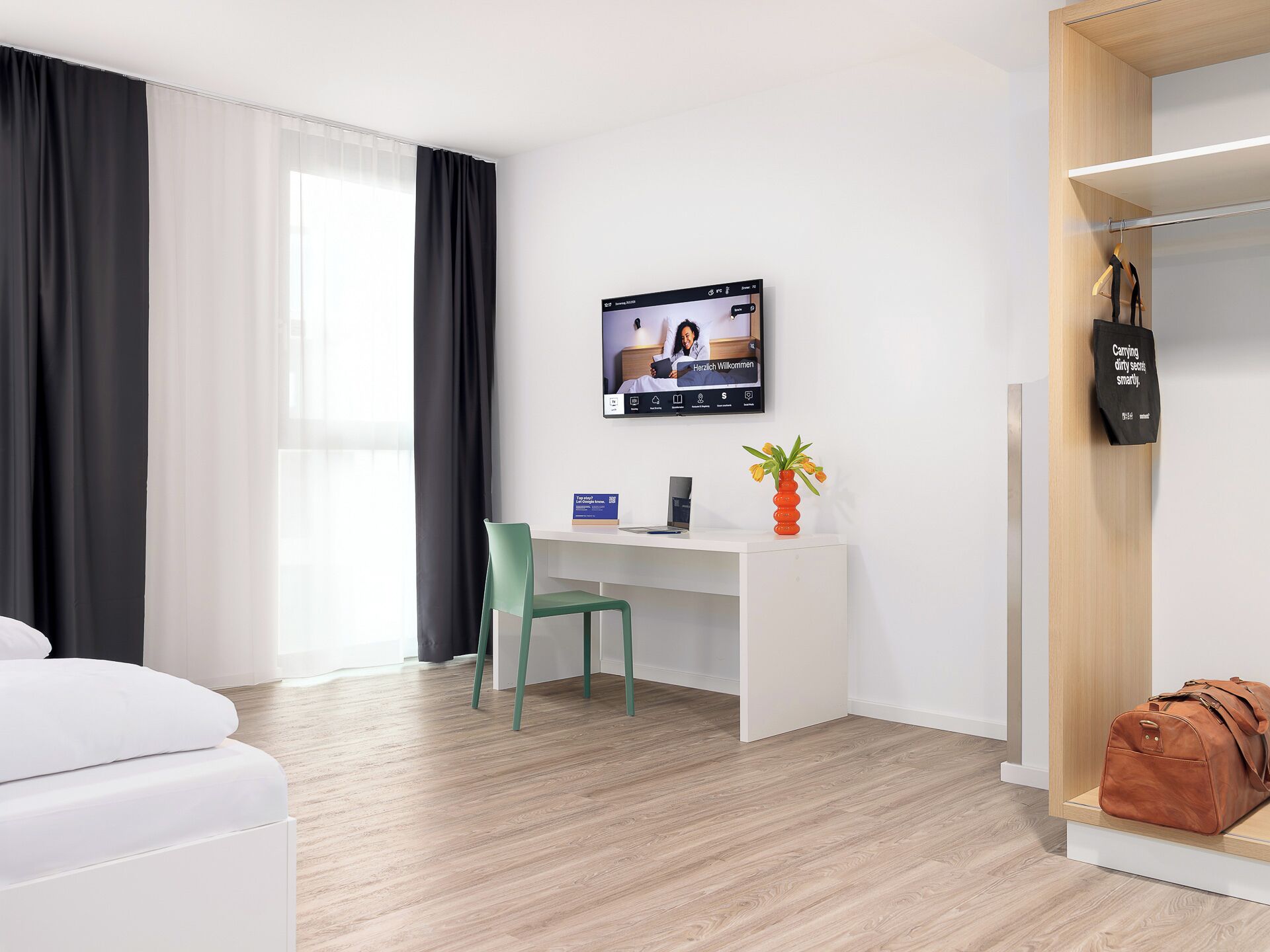 Een hotelkamer met een bureau, een stoel, een bed, een televisiescherm, een kapstok en een tas.
