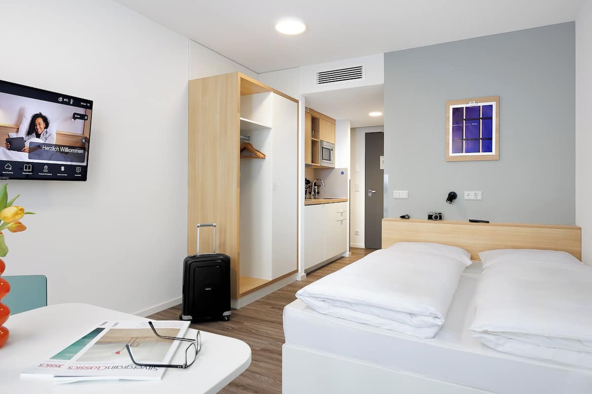 Une chambre d’hôtel comprenant un lit, un bureau avec un ordinateur portable, une valise, une armoire et une petite kitchenette.