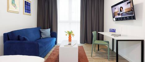 A modern hotel room with a blue sofa, a white desk, a TV, and a large window with curtains.