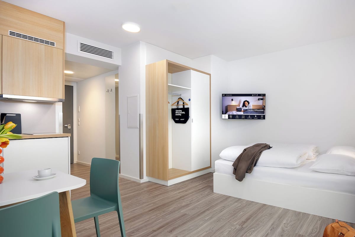 Une chambre d’hôtel moderne équipée d’une kitchenette, d’un téléviseur fixé au mur, d’un canapé et d’une porte en bois.