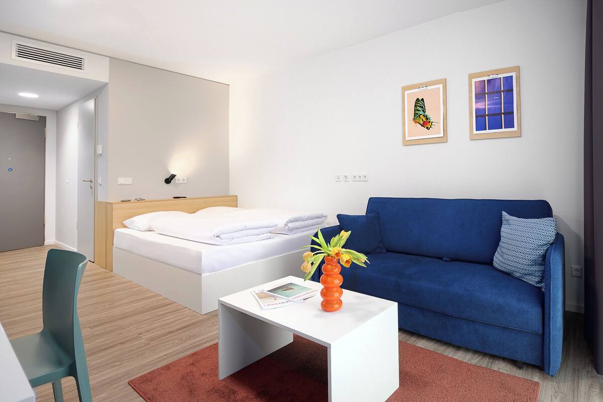 Une chambre d’hôtel moderne avec un lit, un canapé bleu, une table basse blanche ornée d’un vase et un tapis rouge.