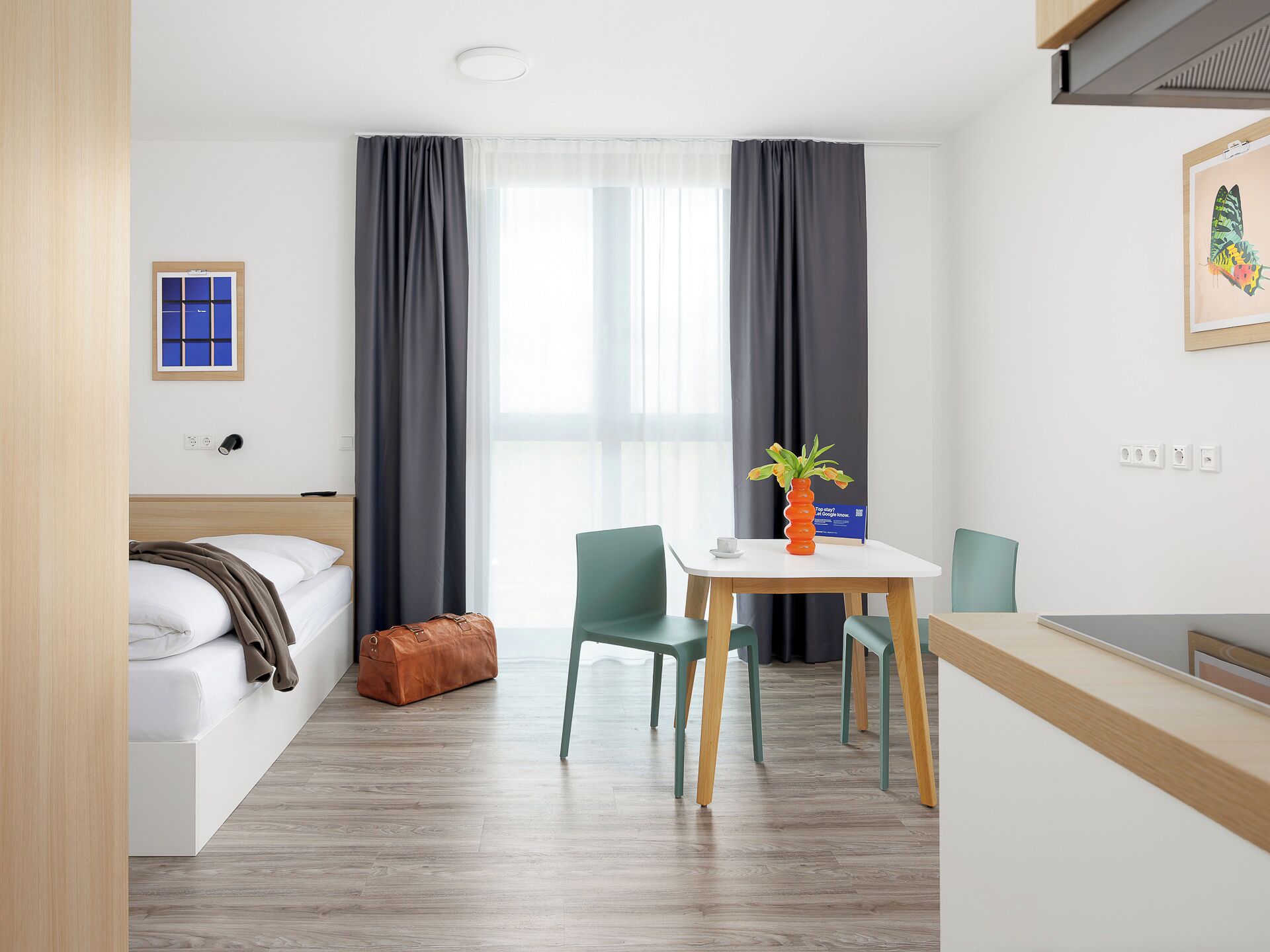 A hotel room with a bed, a desk, a chair, a small table with a vase, and a window with curtains.