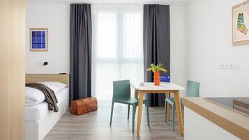 A hotel room with a bed, a desk, a chair, a small table with a vase, and a window with curtains.