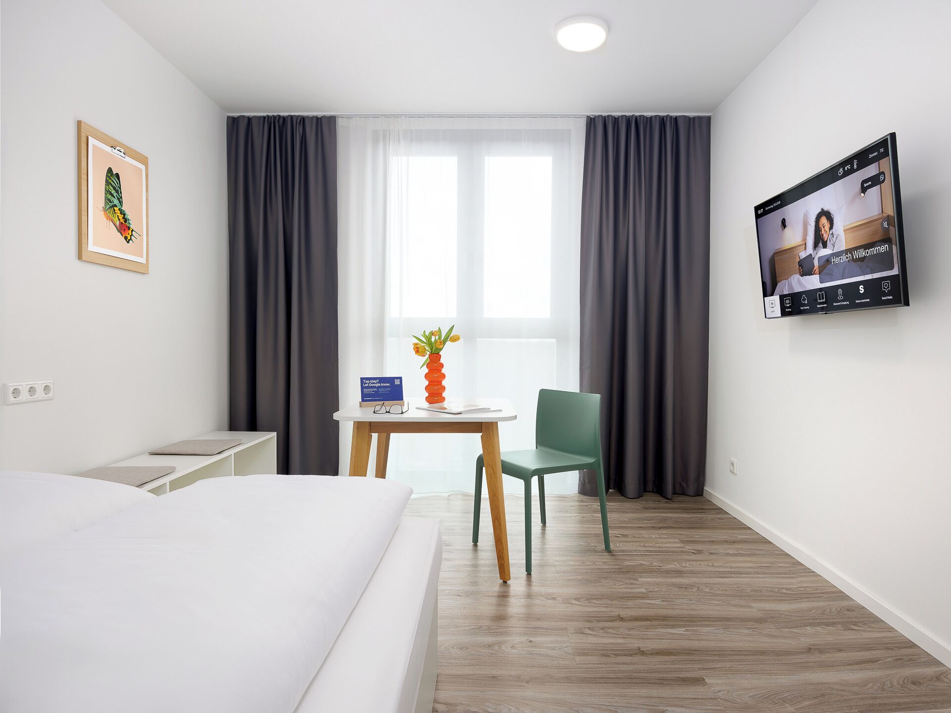 A hotel room with a bed, a desk with a vase of flowers, a chair, a TV on the wall, and a window with curtains.