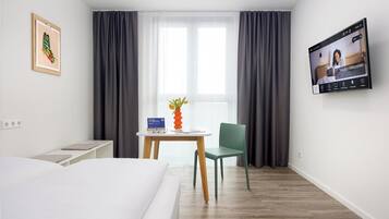 A hotel room with a bed, a desk with a vase of flowers, a chair, a TV on the wall, and a window with curtains.