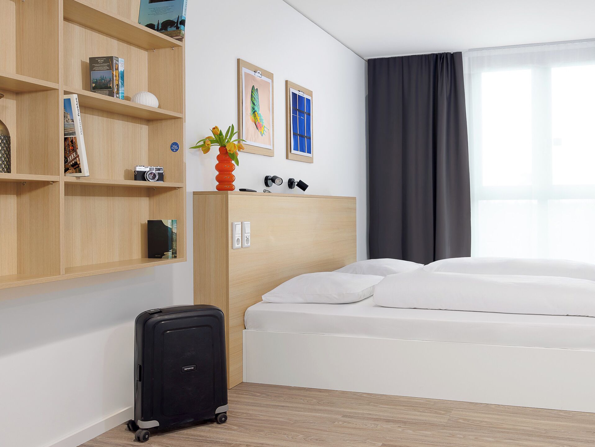 A hotel room with a bed, a wooden shelf with books and a camera, a vase with flowers, and a window with curtains.