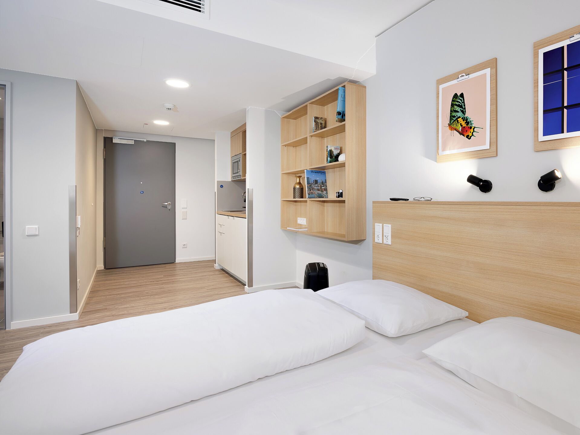 A modern hotel room with a bed, a kitchenette, a door leading to another room, and a wall-mounted shelf with books and a picture.