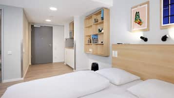A modern hotel room with a bed, a kitchenette, a door leading to another room, and a wall-mounted shelf with books and a picture.