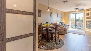 Un coin repas avec une table et des chaises en bois, un ventilateur de plafond et une vue sur l’extérieur grâce à de grandes fenêtres.