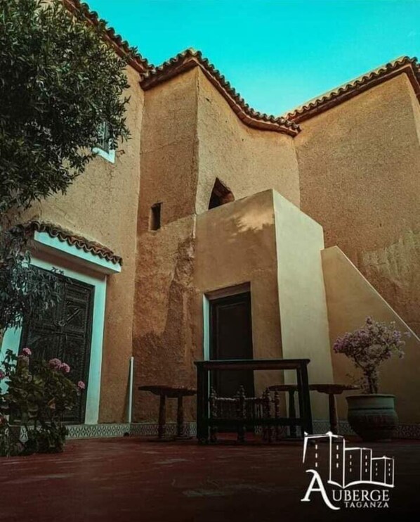 A building with a traditional architectural style, featuring a flat roof and terracotta tiles. There is a door and a window visible, and a bench is placed in front of the building. - Auberge tagnza (Aoulouz)