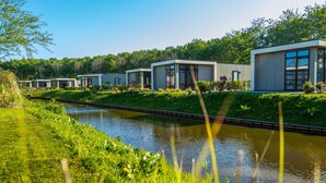 A row of modern, single-story buildings with large windows, situated beside a calm waterway, surrounded by lush greenery. - Modern Holiday Home Near the Golf Course (Velsen-Zuid)