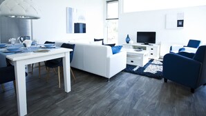 A modern living room with a white sofa, blue armchairs, a dining table set for four, and a flat-screen TV. - Modern Holiday Home Near the Golf Course (Velsen-Zuid)
