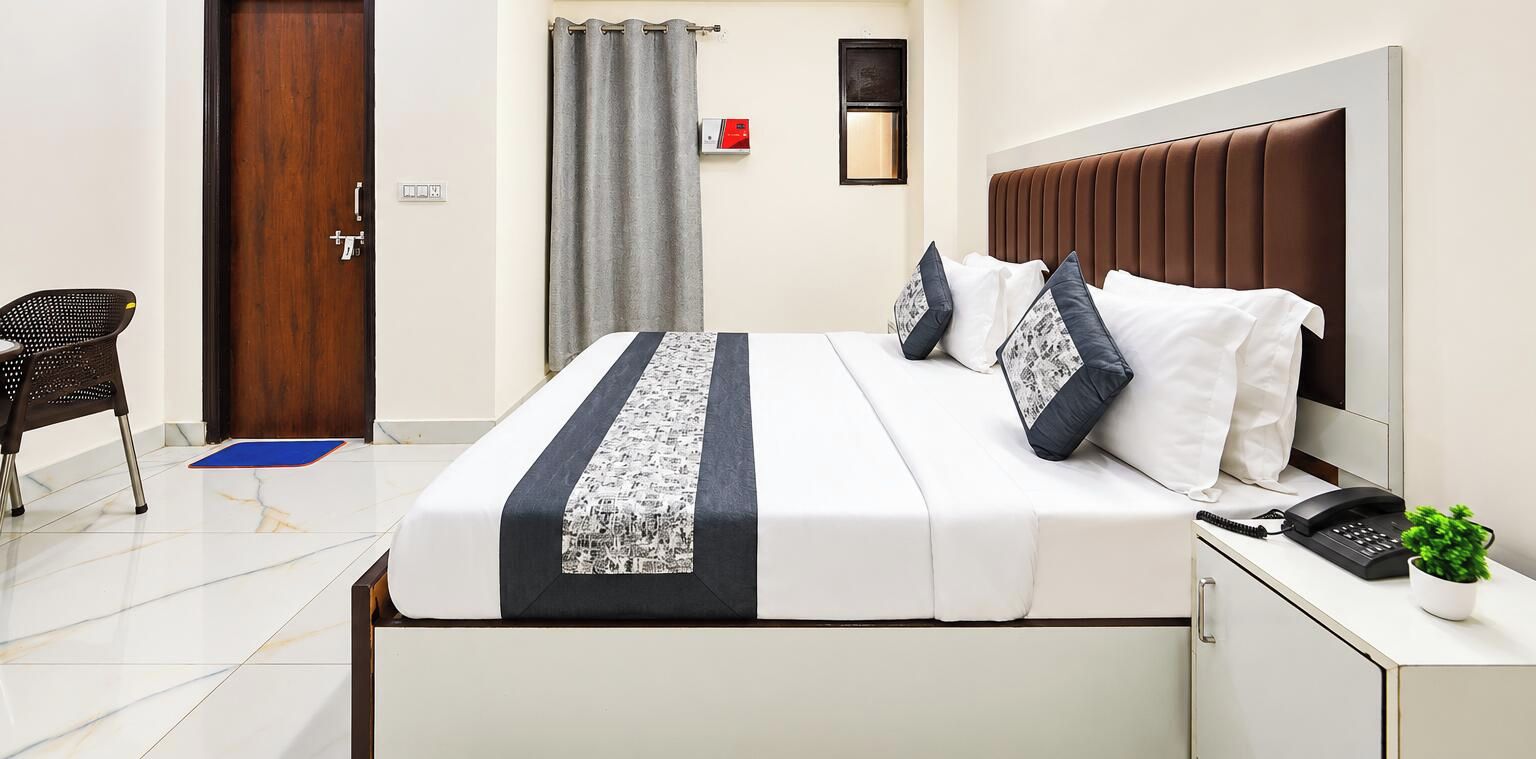 A hotel room with a bed, a desk with a phone and a plant, a chair, a door, and a curtain.