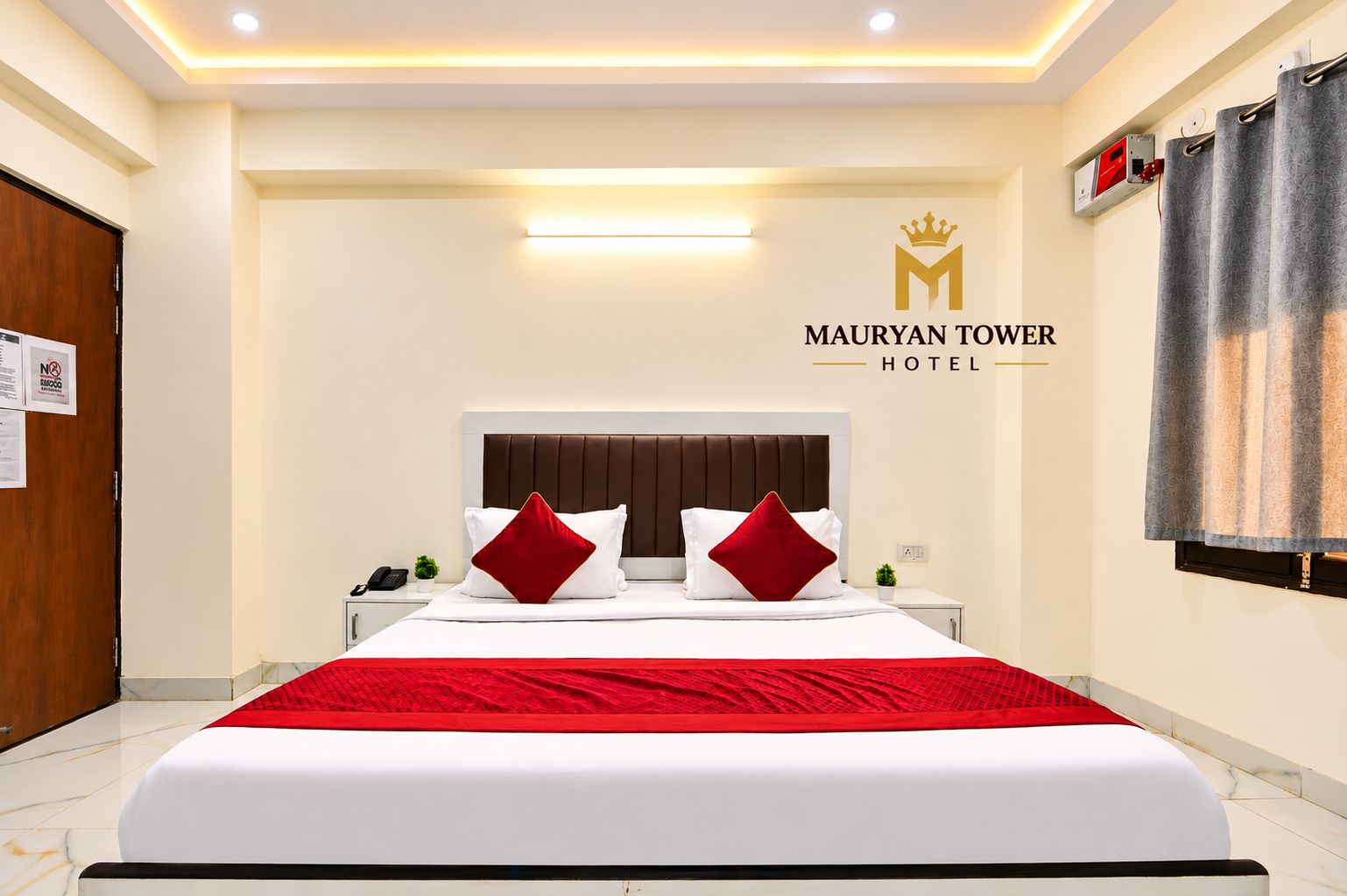 A hotel room with a large bed, a headboard, a nightstand, a phone, a red throw blanket, and a sign with the hotel name.