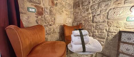 Una habitación de esquina con pared de piedra, un par de sillas naranjas, una mesita con toallas y un mueble decorativo.