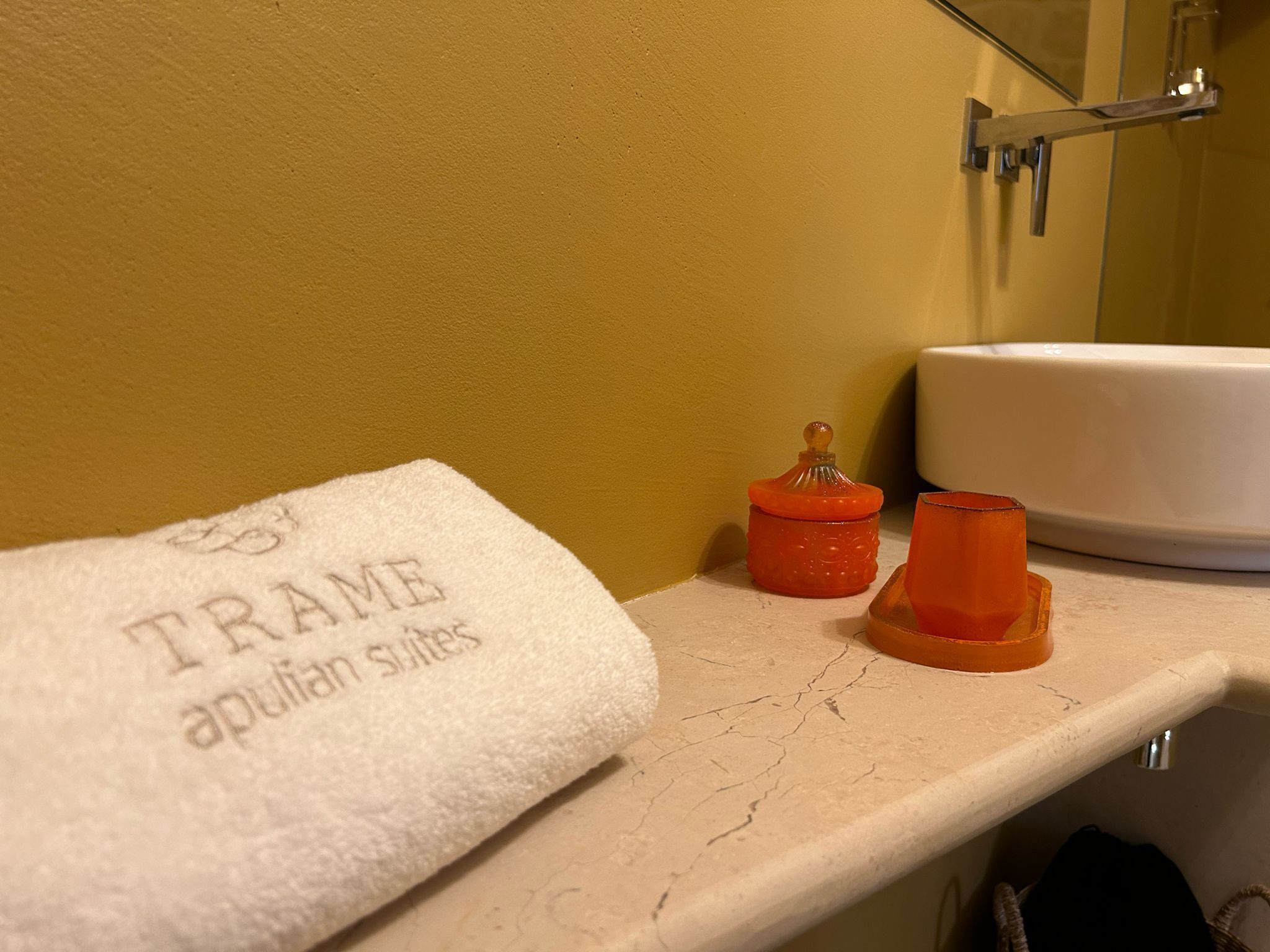 A bathroom with a towel labeled "TRAME apulian suites" on a shelf, a white sink, and two orange containers.