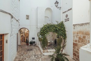 A narrow courtyard with white walls, a stone floor, and a covered seating area with plants.