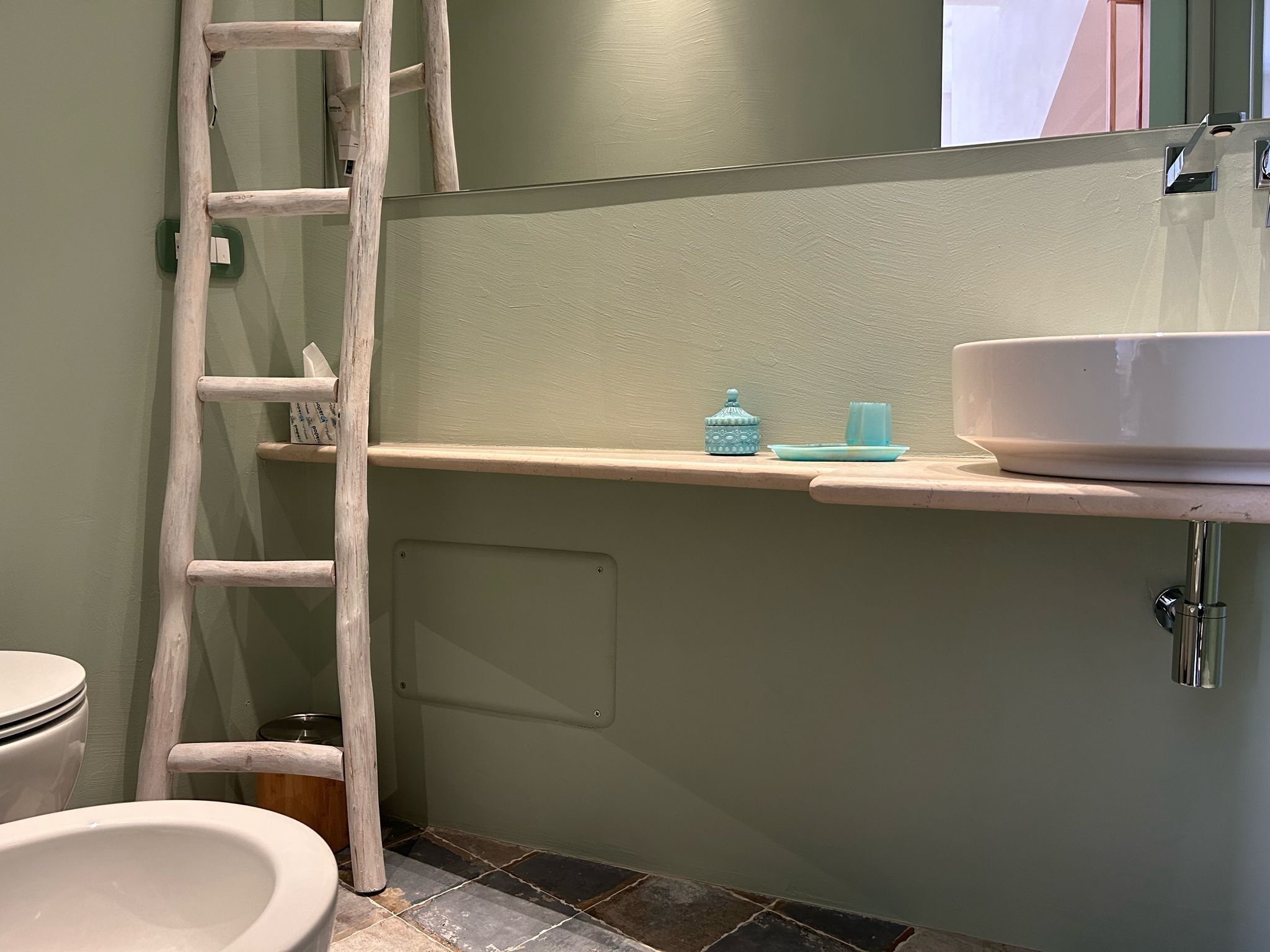 Un baño con un inodoro blanco, un lavamanos blanco con un recipiente grande, una escalera de madera apoyada en la pared y un pequeño estante con un recipiente azul y un vaso.