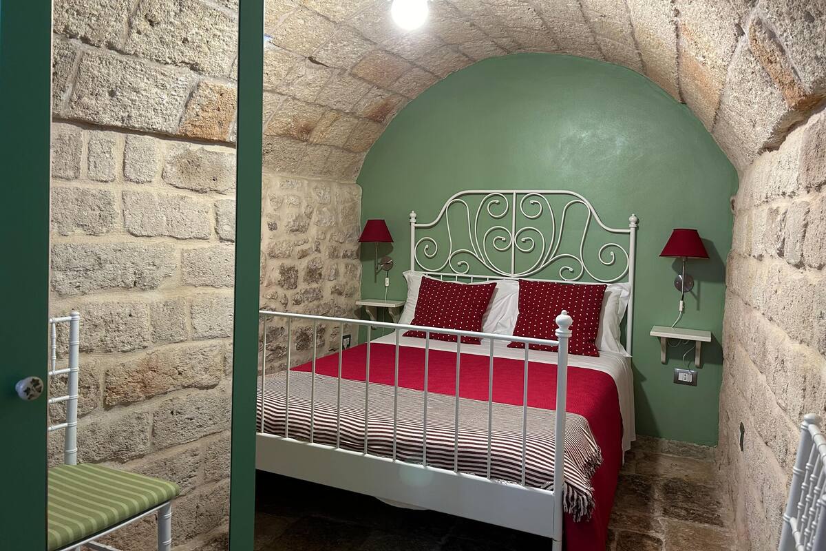 A room with a stone wall, a bed with a white metal headboard, red bedding, and two wall-mounted lamps.