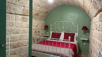 A room with a stone wall, a bed with a white metal headboard, red bedding, and two wall-mounted lamps.