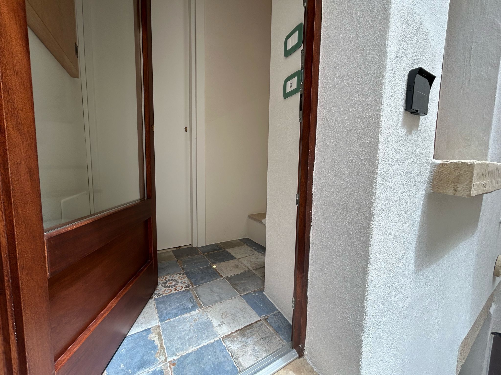 Een gang met een betegelde vloer, een houten deur en een witte muur met een klein groen bordje.