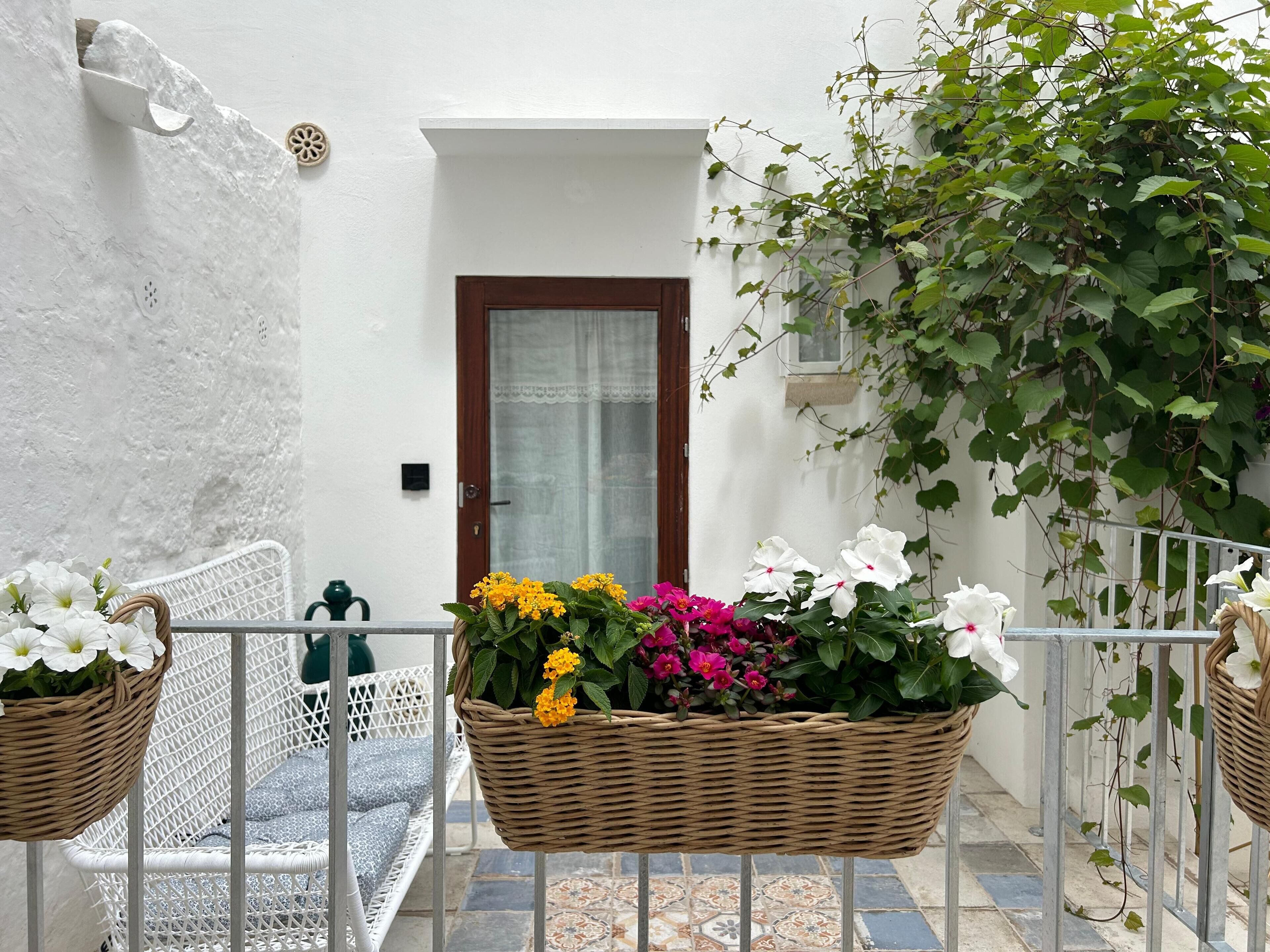 Un balcón con cestas de mimbre llenas de flores, una silla blanca y una puerta al fondo.