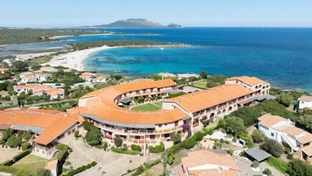 An aerial view of a coastal hotel with a curved building, beachfront access, and a clear view of the sea and distant mountains. - Mishti Blu Studio al Mare by Wonderful Italy (Olbia)
