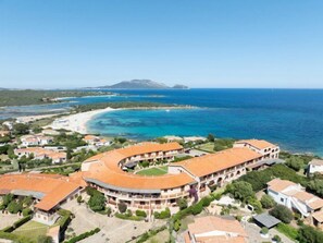 An aerial view of a coastal hotel with a curved building, beachfront access, and a clear view of the sea and distant mountains. - Mishti Blu Studio al Mare by Wonderful Italy (Olbia)