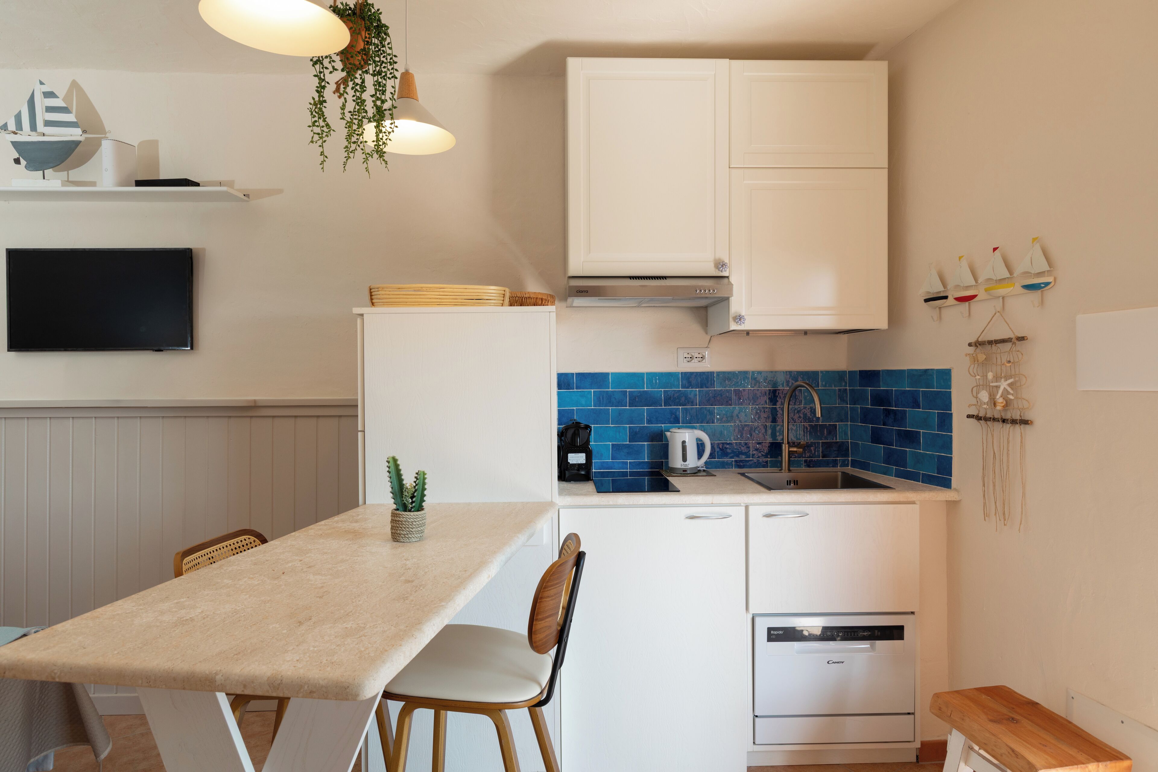 A compact kitchen with white cabinets, a blue-tiled backsplash, a built-in dishwasher, and a dining area with a table and chairs.