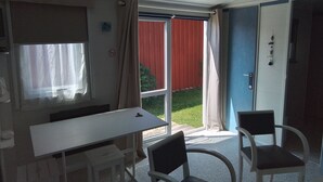A room with a white table and two chairs, a window with curtains, and a sliding glass door leading to a red fence and green lawn. - Mobilhome en Presquîle Guerandaise Pour 4 Personnes (Saint-Molf)