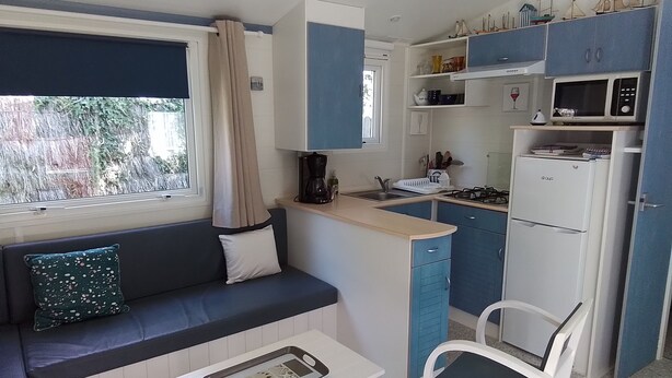 A compact kitchenette with a stove, microwave, and sink. There is a small dining area with a table and chair. A window with curtains is visible, and the room has a blue and white color scheme. - Mobilhome en Presquîle Guerandaise Pour 4 Personnes (Saint-Molf)
