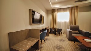 Quarto de hotel com televisão na parede, área de estar com sofá e poltronas, uma pequena mesa e um tapete estampado.