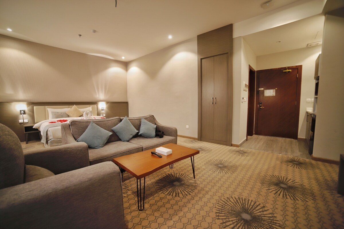 A hotel room with a sofa, a wooden coffee table, a patterned carpet, a wardrobe, and a door leading to a bathroom.