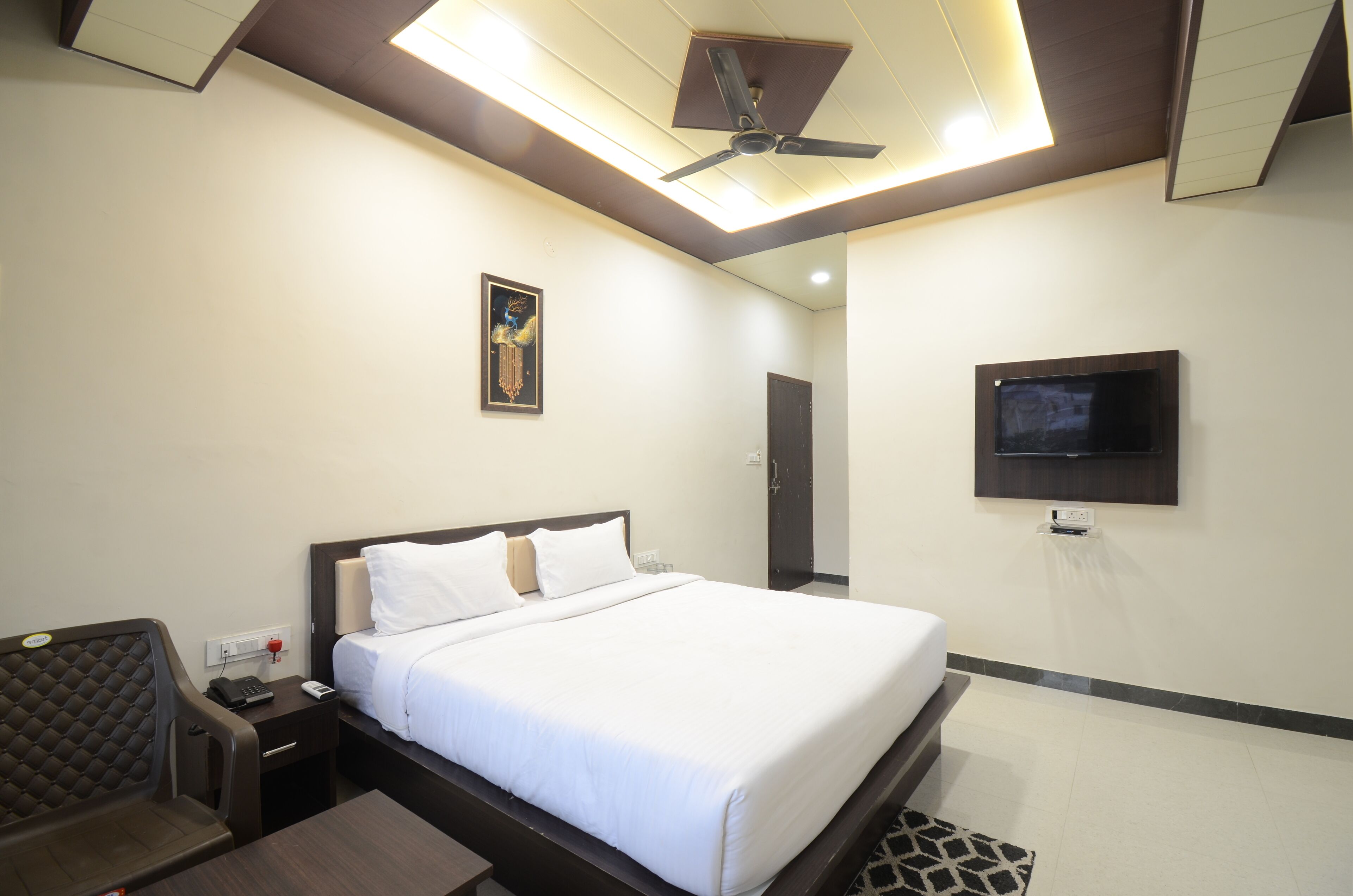 Habitación de hotel con cama, mesita de noche, silla, ventilador de techo, obra de arte, televisión y puerta.