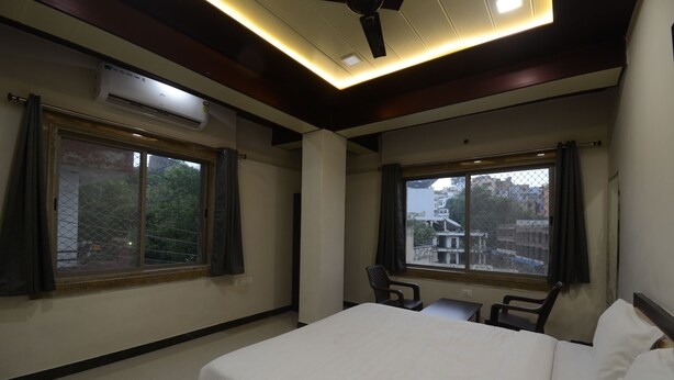 A hotel room with a bed, two chairs, a desk, and large windows with curtains. - Hotel Shri Dhwaja Inn (Nathdwara)
