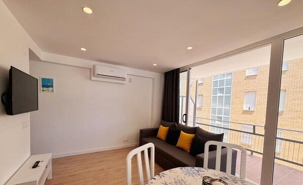 A modern living room with a sofa, a TV mounted on the wall, and a balcony with a view of the city. - Beachside Apartment with Free WiFi, Near Nightlife & Port Aventura, Sleeps 6 (Salou)