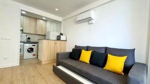 A modern living room with a grey sofa, two black and one yellow cushion, a washing machine, and a kitchen area with wooden cabinets and a microwave. - Charming Apartment Steps from Capellans Beach – Free WiFi, Fully Equipped Kitchen, Sleeps 4 (Salou)