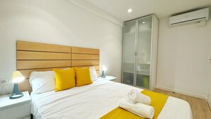A hotel room with a wooden headboard, a bed with white and yellow bedding, a nightstand with a lamp, a wardrobe, and an air conditioning unit. - Charming Apartment Steps from Capellans Beach – Free WiFi, Fully Equipped Kitchen, Sleeps 4 (Salou)