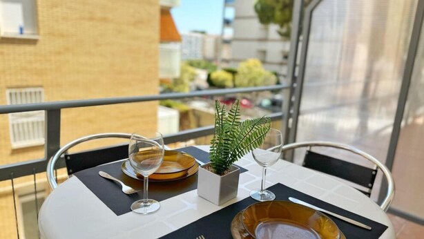 A table set for two with a small potted plant, two wine glasses, and a bowl of fruit on a balcony with a view of buildings and trees. - Charming Apartment Steps from Capellans Beach – Free WiFi, Fully Equipped Kitchen, Sleeps 4 (Salou)