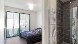 Ein Schlafzimmer mit einem Bett, einer Schiebetür zu einem Balkon sowie einer Dusche mit dunkelgefliesten Wänden und einer Duschvorhang.