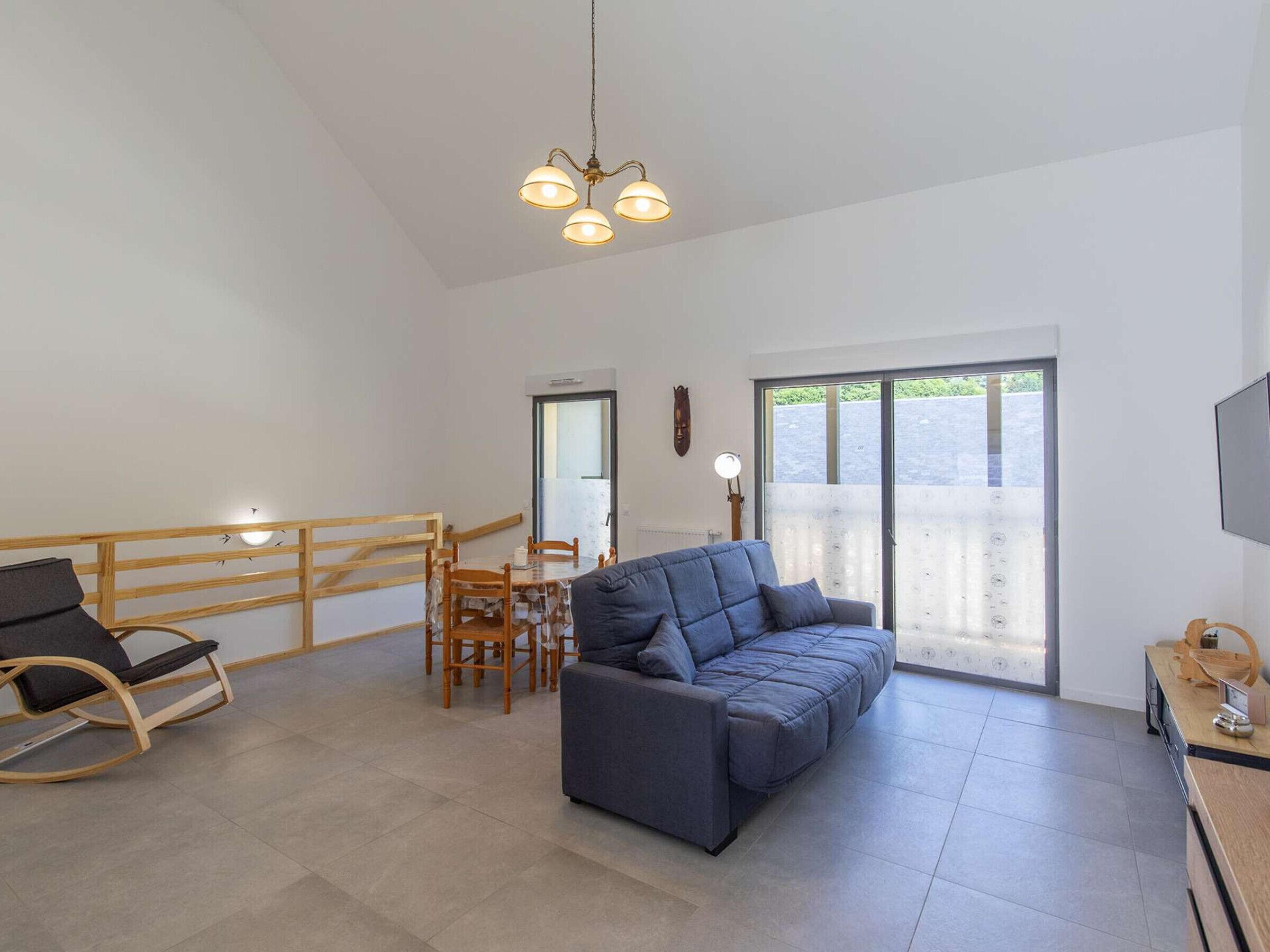A spacious living area with a sofa, a dining table with chairs, a wooden rocking chair, and a balcony with a view.