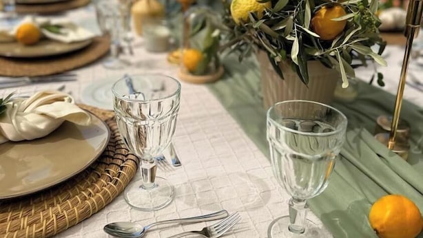 Mesa lista para una comida, con vasos transparentes, platos y limones, con una montaña y una piscina de fondo.
