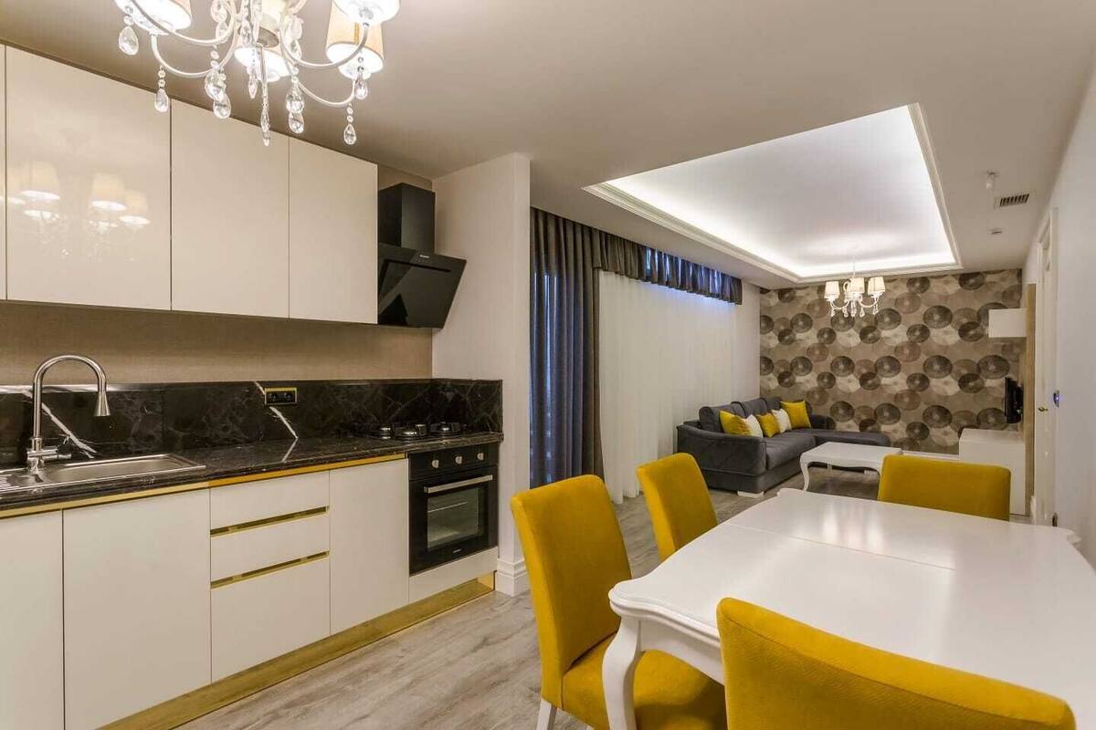 A modern kitchen with a black countertop, white cabinets, and a built-in oven. There is a dining area with a white table and yellow chairs.